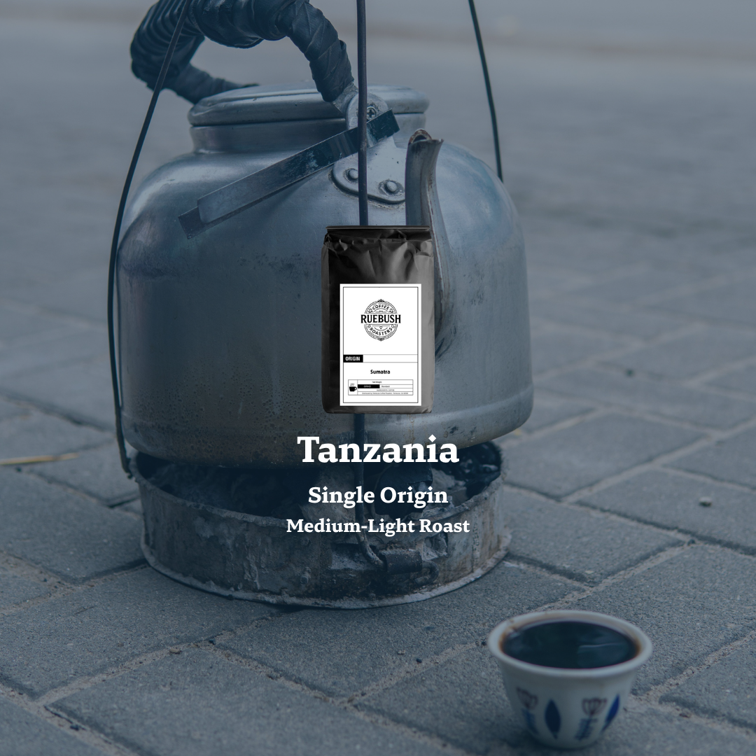 Metal coffee pot with a bag of coffee labeled Tanzania Single Origin on a stone pavement.